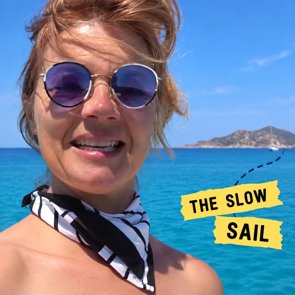 Close portrait of Noemi wearing sunglasses and a neck scarf, with The Slow Sail stickers beside the sea.