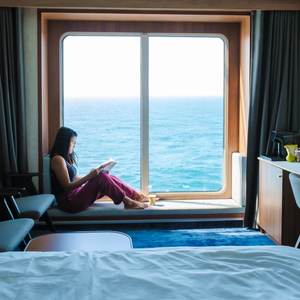 A passenger reading beside a large cabin window at sea