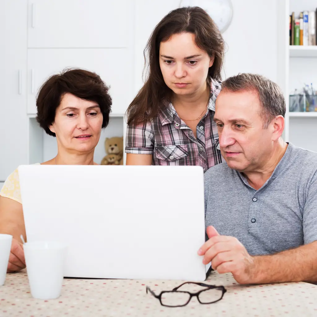 An older couple and their adult daughter studying a laptop together at a kitchen table
