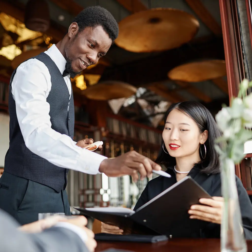 A waiter pointing out a dish on the menu to a seated guest